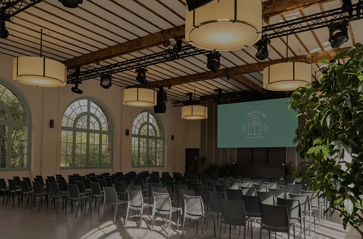 Grande salle de conférence élégante avec poutres apparentes, luminaires suspendus, rangées de chaises face à un écran de projection, baignée de lumière naturelle.